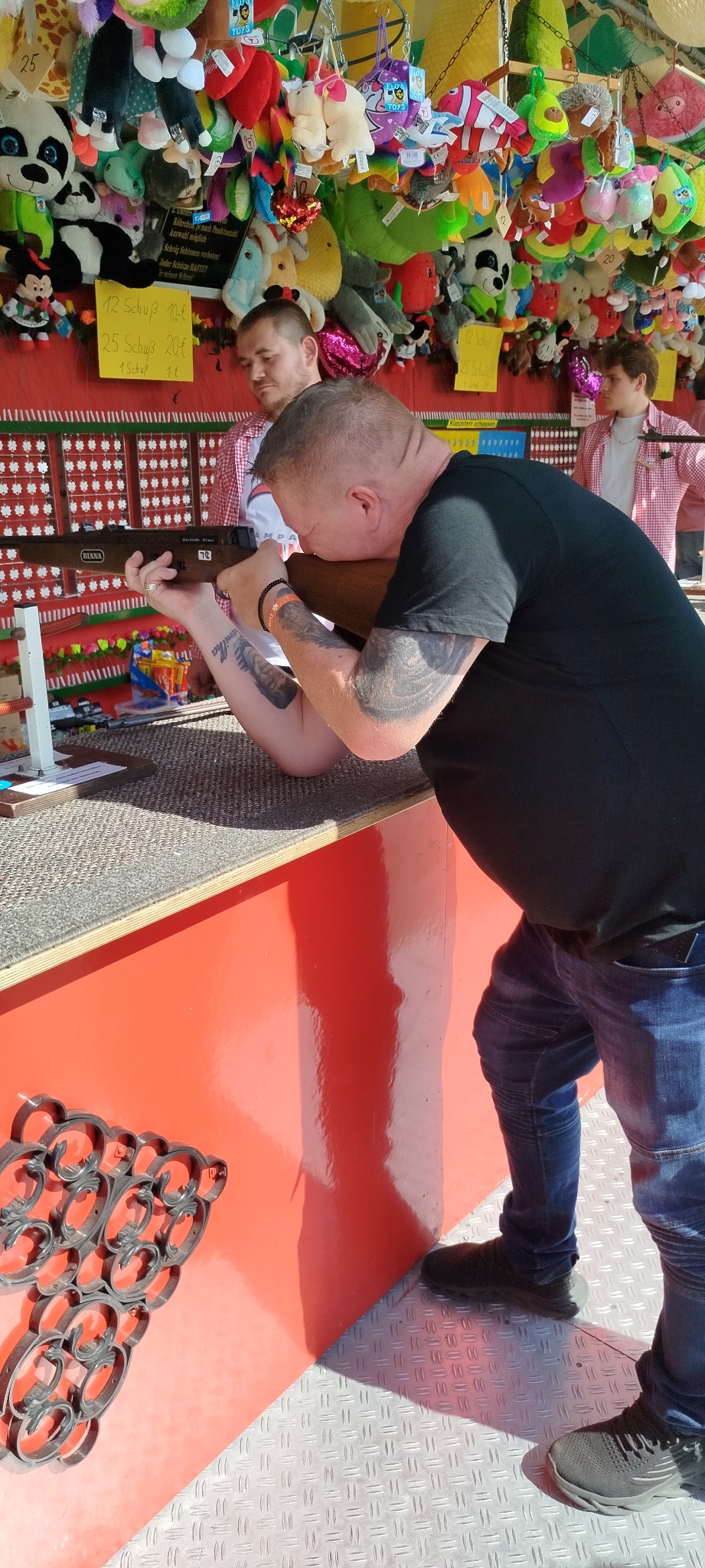 Mężczyzna z tatuażami celuje z wiatrówki do tarczy na strzelnicy, w tle wiszące pluszowe zabawki.