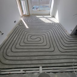 F.U.H ALFA - Montaż ogrzewania podłogowego w szarej wylewce, widoczne pętle rur, okno dachowe z widokiem na budynek i samochód dostawczy.