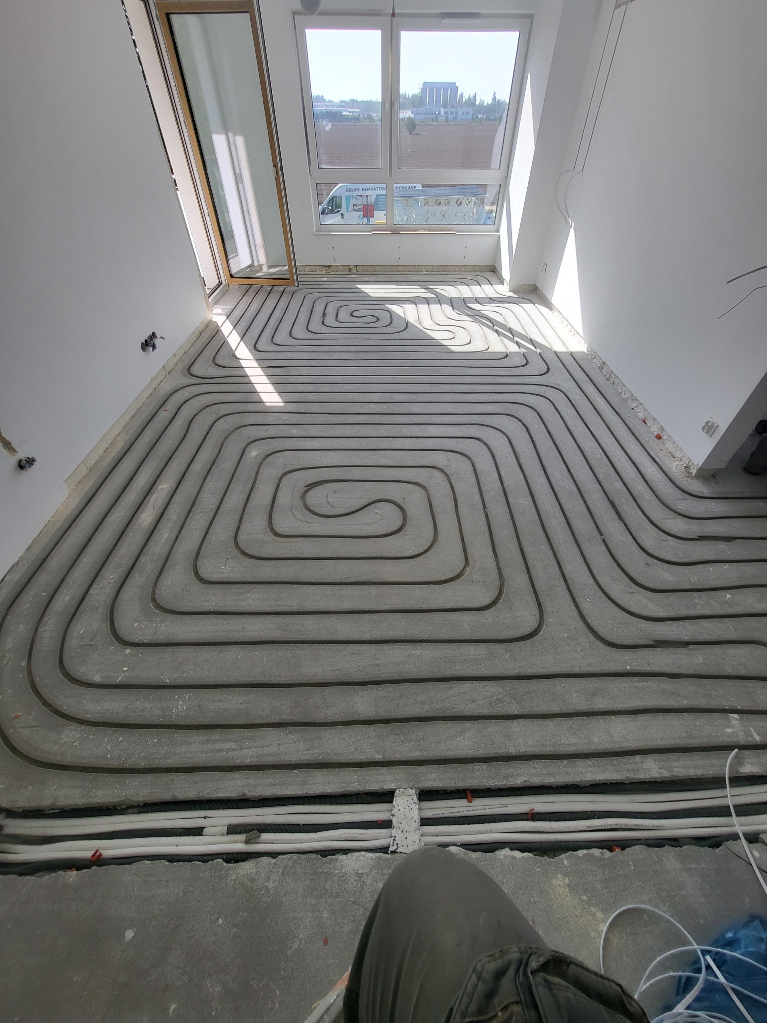 Montaż ogrzewania podłogowego w szarej wylewce, widoczne pętle rur, okno dachowe z widokiem na budynek i samochód dostawczy.