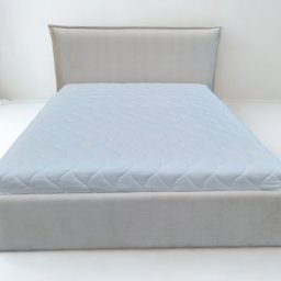 Fotello Meble Robert Śmiałek - Tapicerowane łóżko z wezgłowiem w jasnej tonacji, pokryte błękitnym materacem z pikowaniem, na białym tle. Minimalistyczny design, stonowana kolorystyka.