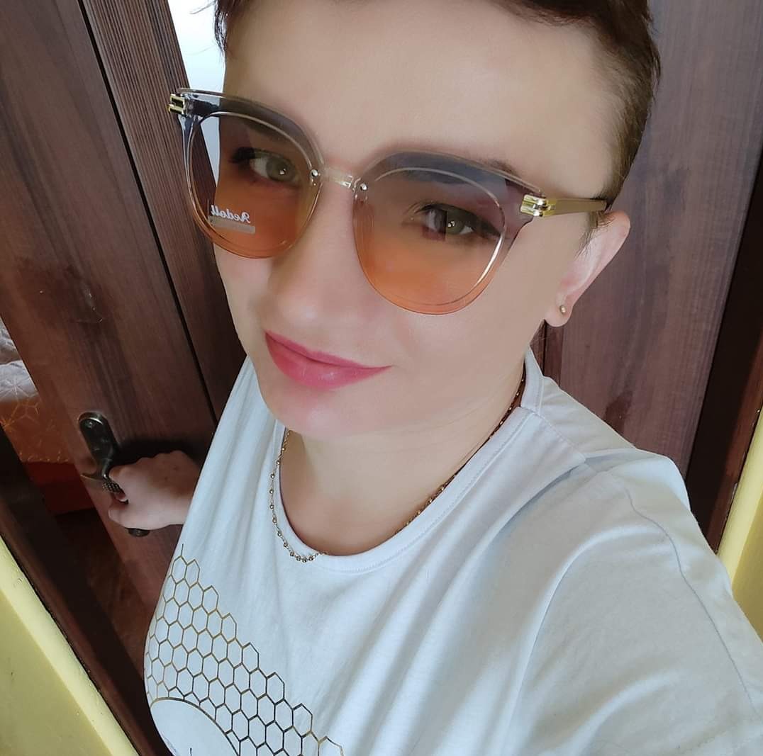 Portret kobiety w okularach z brązowymi szkłami, uśmiechniętej, stojącej przy drewnianych drzwiach z klamką, ubrana w białą koszulkę z geometrycznym wzorem.