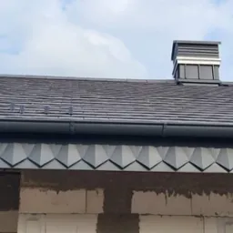 Szary dach z dachówki ceramicznej z nowoczesnym kominem i rynną, widoczne obróbki blacharskie na krawędzi dachu, ściana budynku w trakcie budowy.