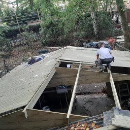 Jakub Wiencko - Pracownik w szarej koszulce i spodniach roboczych montuje drewnianą konstrukcję dachu na tle zalesionego terenu, widoczne elementy konstrukcyjne i narzędzia pracy.