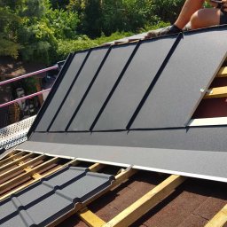 Jakub Wiencko - Układanie szarej blachy na dachu z widoczną konstrukcją drewnianą i częścią dekarza z narzędziami, w tle zieleń drzew.