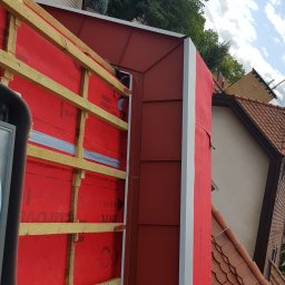 Jakub Wiencko - Konstrukcja dachu z drewnianymi łatami i czerwoną membraną izolacyjną, widok z góry na fragment budynku z dachówką ceramiczną w tle.