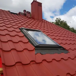 Czerwony dach z blachodachówki modułowej w Krypno Kościelne, z oknem dachowym i kominem w tym samym kolorze, na tle błękitnego nieba z chmurami.