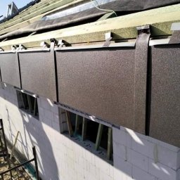 Jakub Wiencko - Montaż paneli elewacyjnych z metalu na dachu budynku w trakcie budowy, widoczne elementy konstrukcji dachu i ściany z białych bloczków.