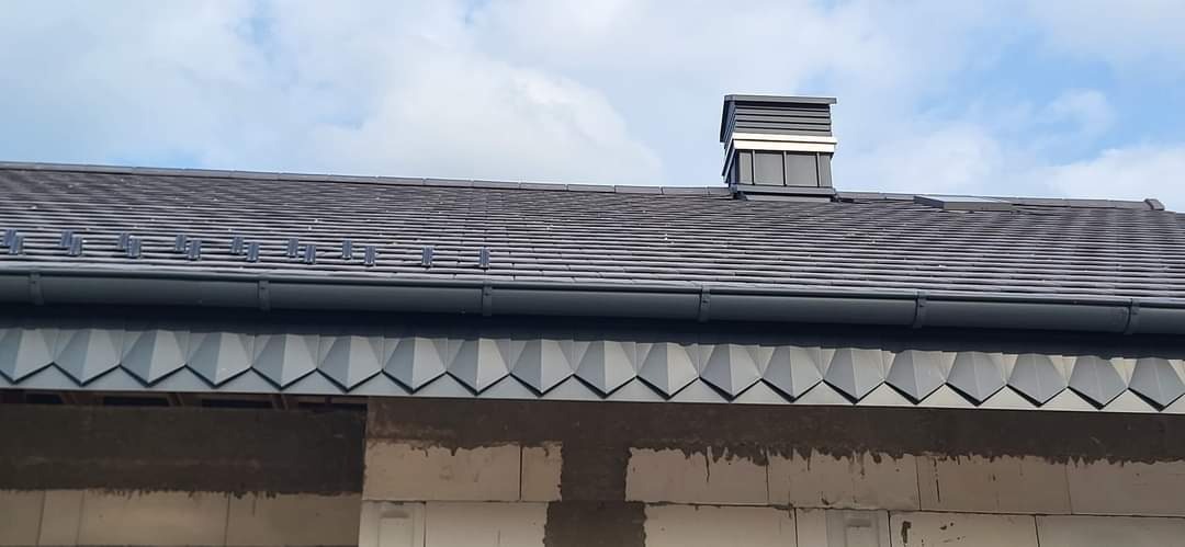 Szary dach z dachówki ceramicznej z nowoczesnym kominem i rynną, widoczne obróbki blacharskie na krawędzi dachu, ściana budynku w trakcie budowy.