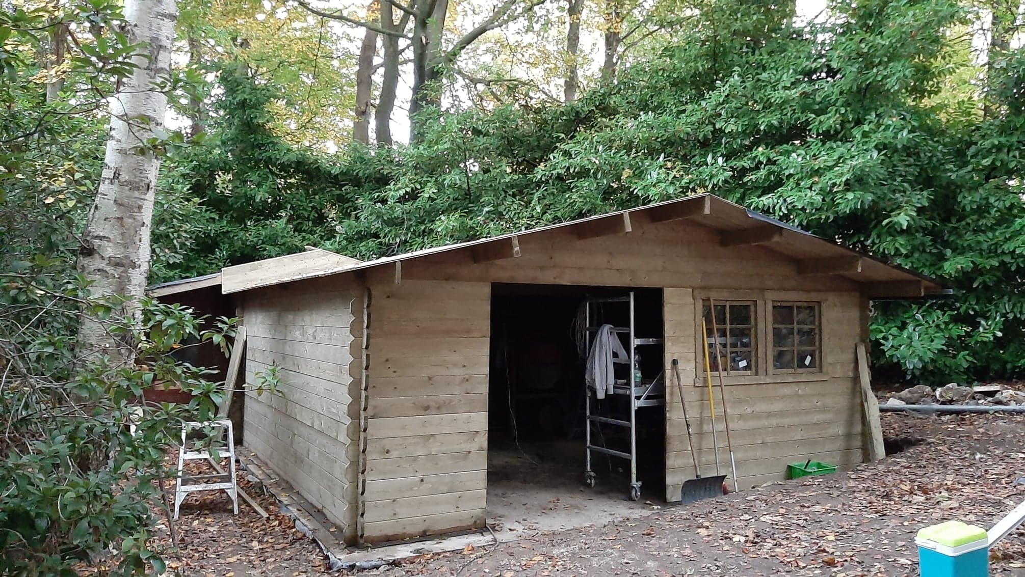 Drewniana konstrukcja garażowa z dwuspadowym dachem, częściowo wykończona, z widoczną drabiną i narzędziami w otoczeniu drzew