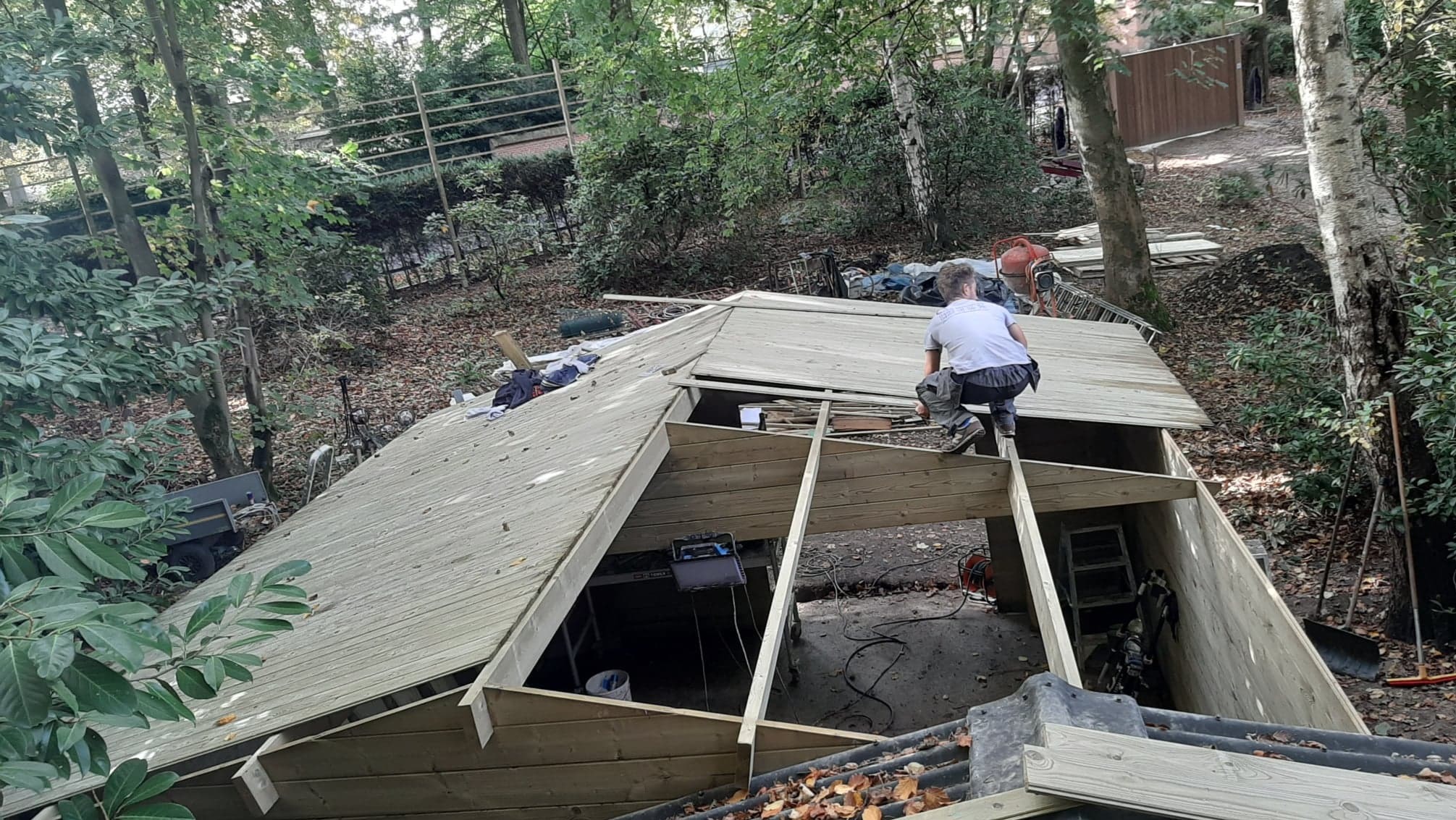 Pracownik w szarej koszulce i spodniach roboczych montuje drewnianą konstrukcję dachu na tle zalesionego terenu, widoczne elementy konstrukcyjne i narzędzia pracy.