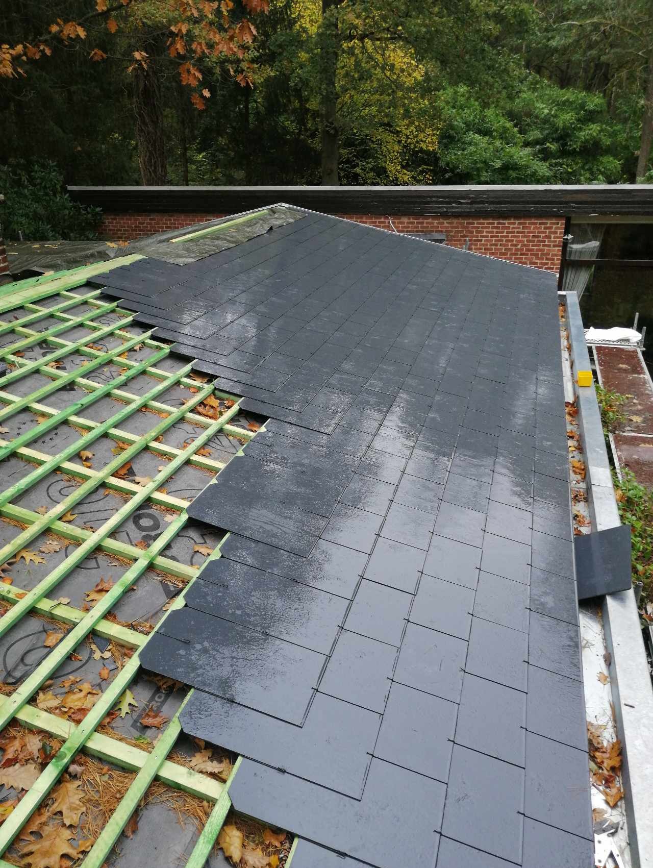 Montaż szarej dachówki na łatach z widoczną membraną dachową, częściowo pokryty dach, liście na dachu.