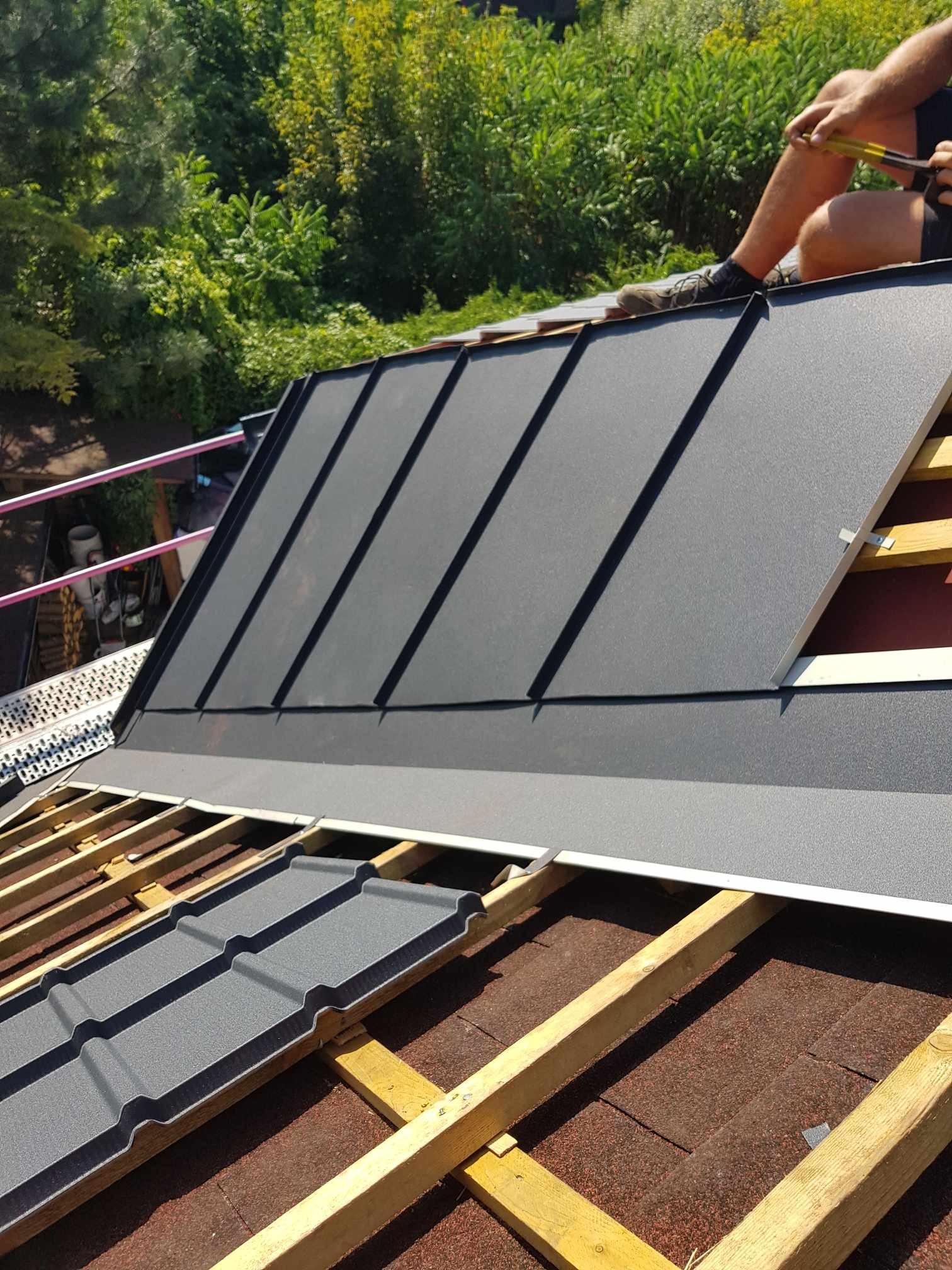 Układanie szarej blachy na dachu z widoczną konstrukcją drewnianą i częścią dekarza z narzędziami, w tle zieleń drzew.