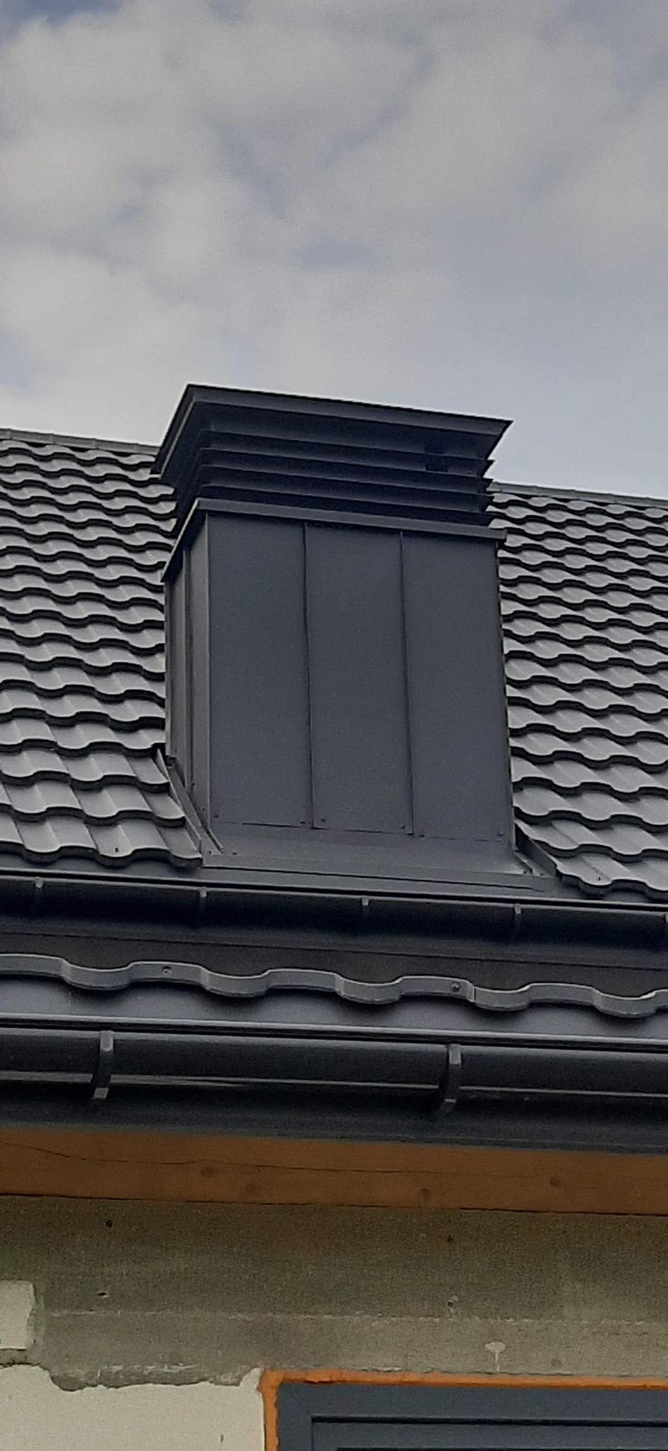 Szary, blaszany komin wentylacyjny na dachu z blachodachówki, z widoczną rynną i fragmentem ściany budynku.