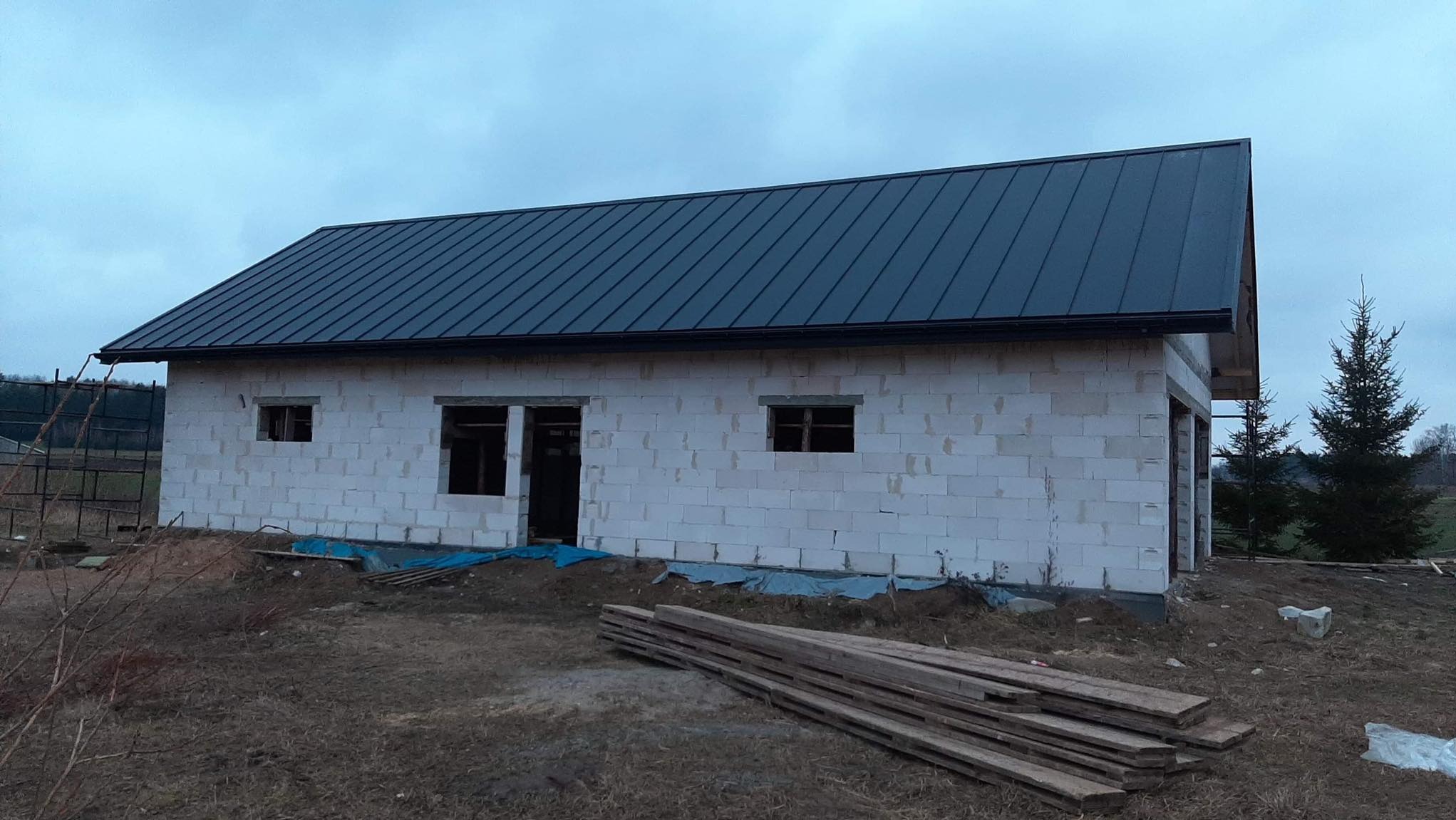 Niedokończony budynek z białego bloczka z ciemnym, metalowym dachem i rynną, widoczne deski na ziemi, pochmurne niebo.