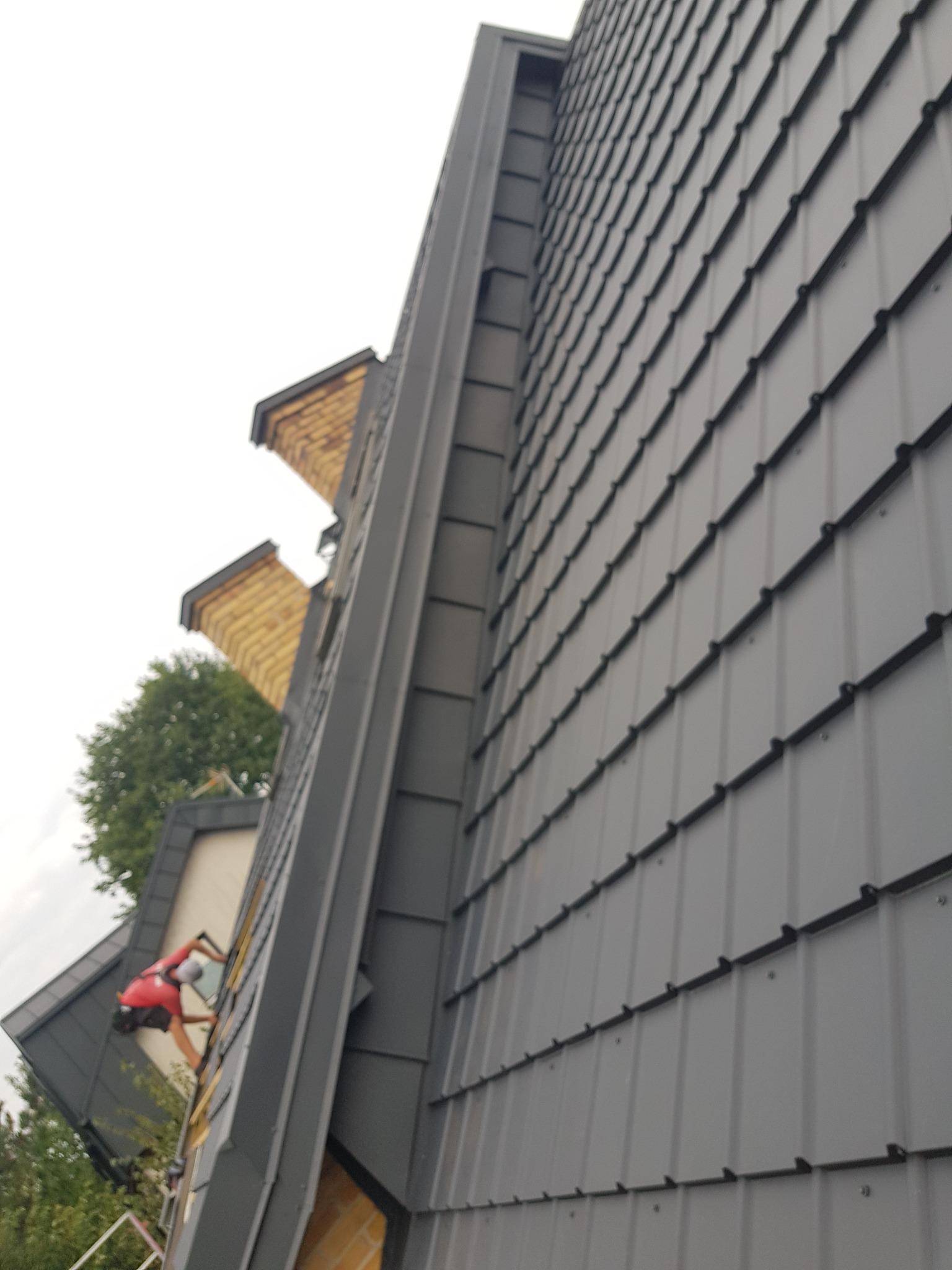 Szary dach z blachy trapezowej z widocznym systemem mocowania, widok z dołu na budynek z ceglanymi elementami i pracownikiem w czerwonej koszulce na drabinie.