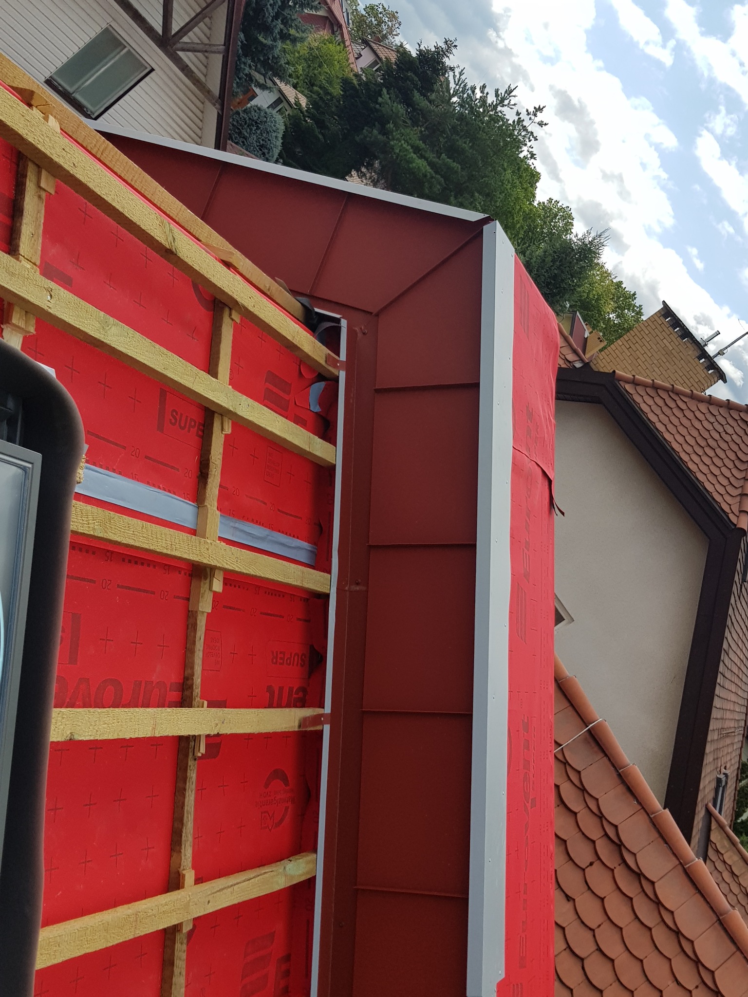 Konstrukcja dachu z drewnianymi łatami i czerwoną membraną izolacyjną, widok z góry na fragment budynku z dachówką ceramiczną w tle.