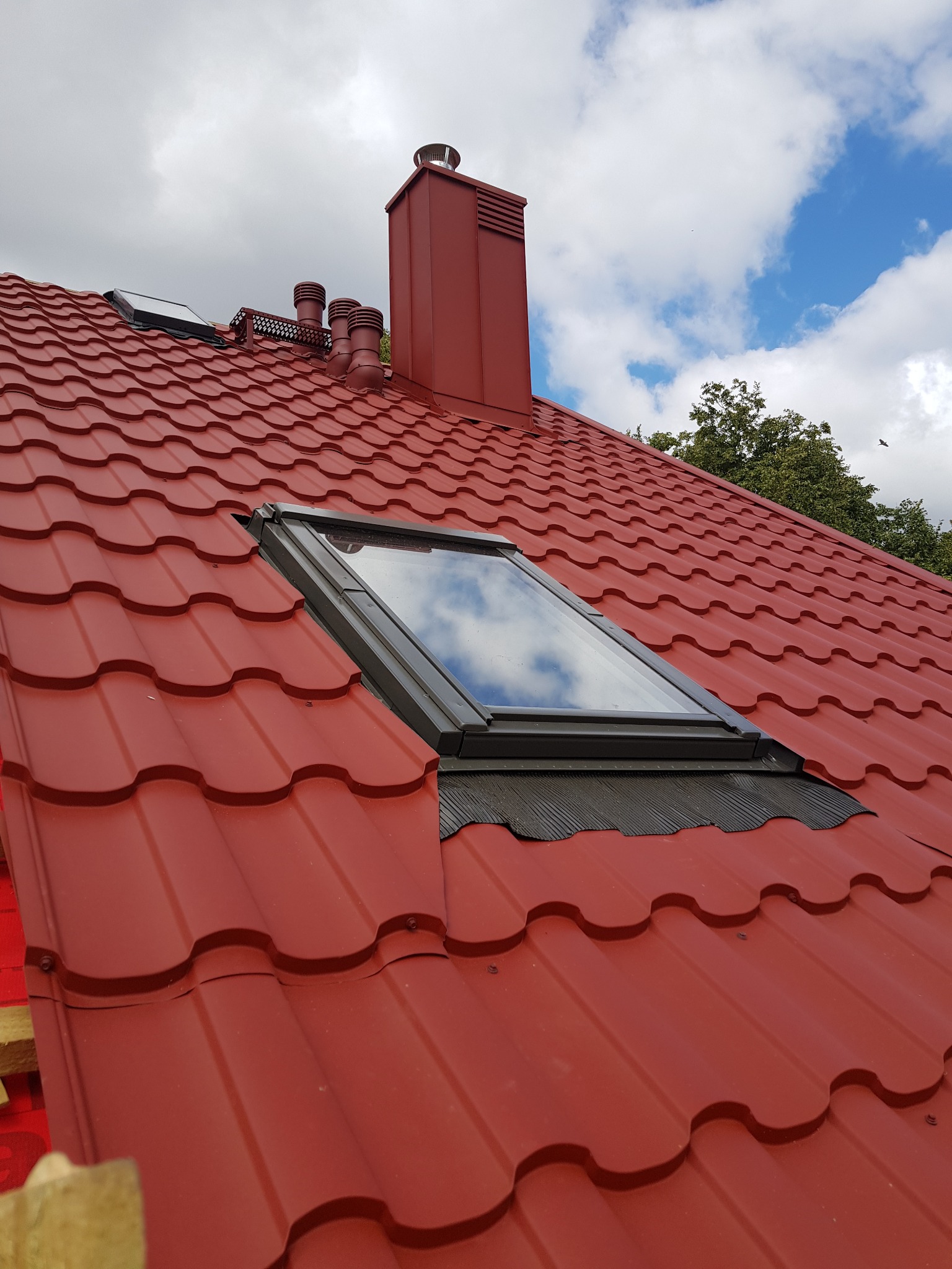 Czerwony dach z blachodachówki modułowej w Krypno Kościelne, z oknem dachowym i kominem w tym samym kolorze, na tle błękitnego nieba z chmurami.