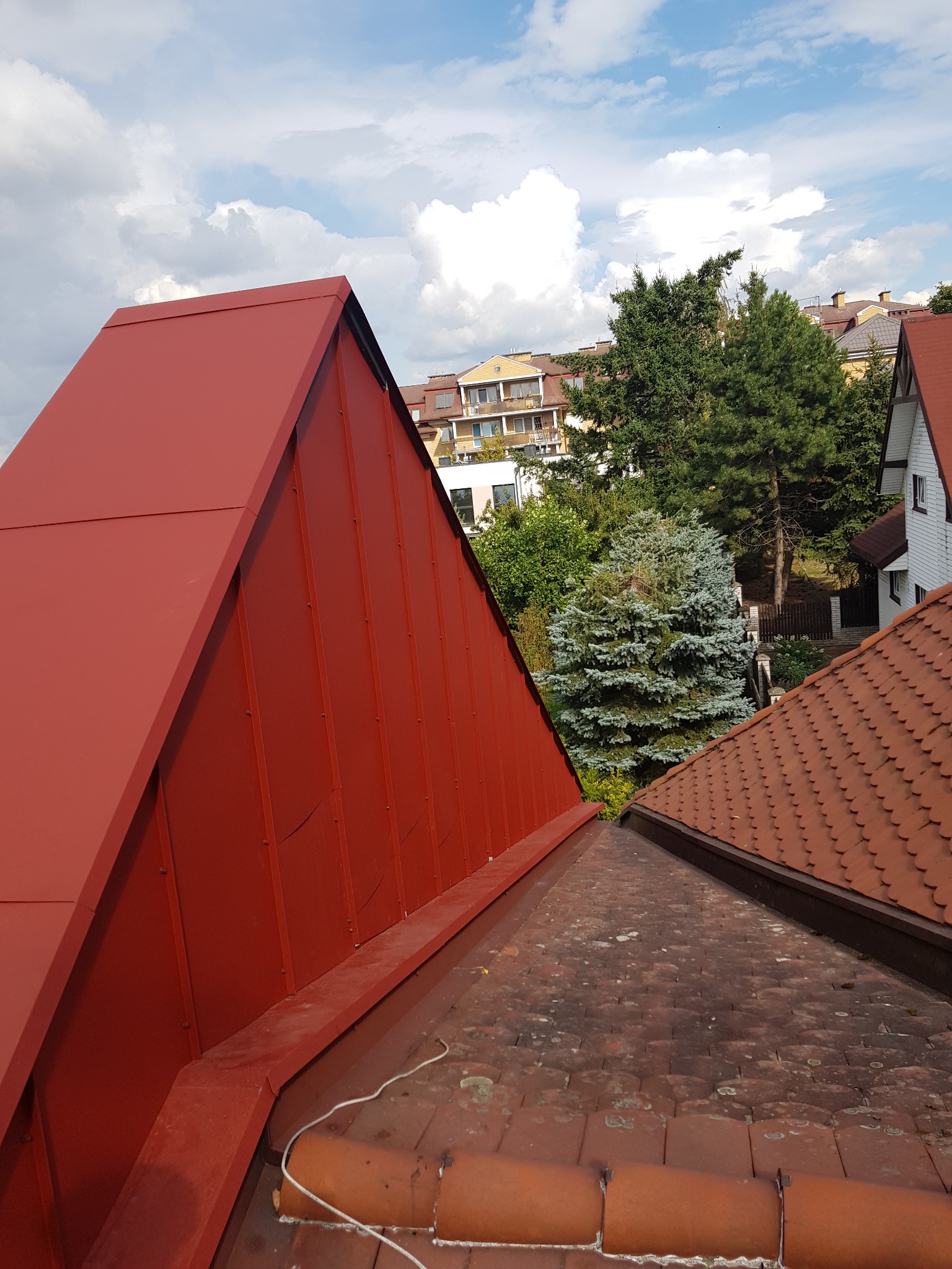 Widok z góry na dwa dachy: jeden z nową, czerwoną blachą trapezową, drugi z dachówką ceramiczną pokrytą mchem, na tle zabudowy miejskiej i błękitnego nieba z białymi chmurami.