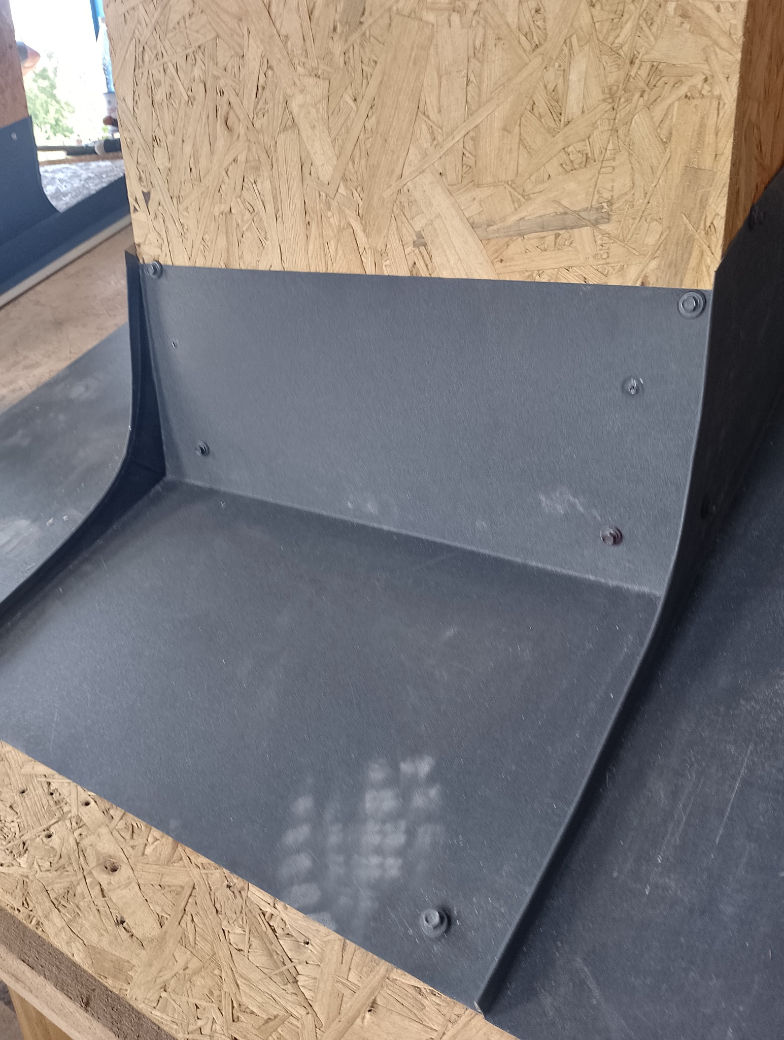 Szara, metalowa obróbka blacharska przymocowana śrubami do drewnianej płyty OSB, widoczne ślady po montażu.