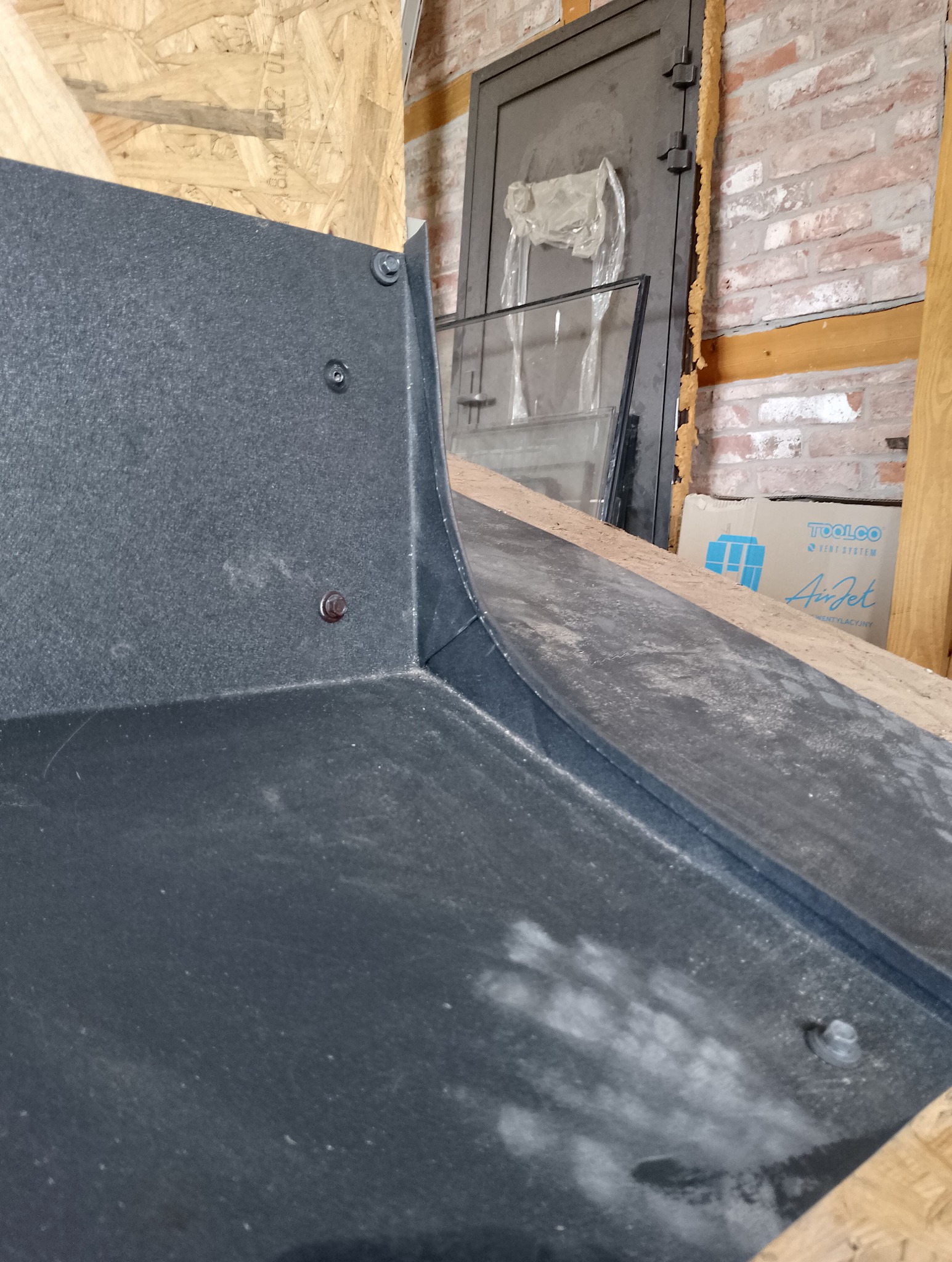 Szara, metalowa obróbka blacharska z widocznymi śrubami mocującymi, umieszczona w surowym wnętrzu z elementami drewnianymi i ceglanymi w tle.