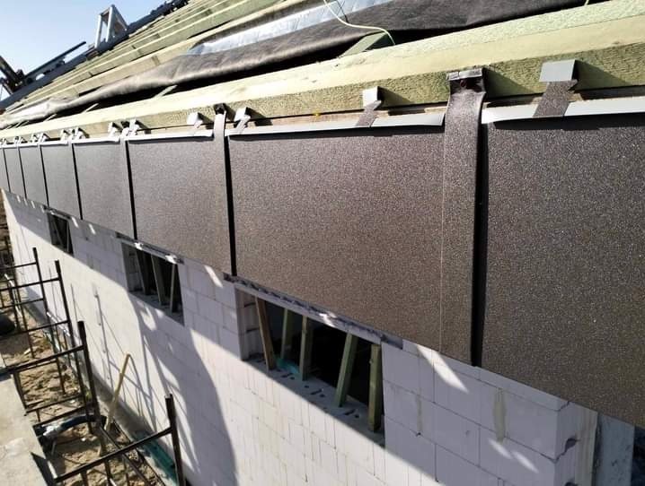 Montaż paneli elewacyjnych z metalu na dachu budynku w trakcie budowy, widoczne elementy konstrukcji dachu i ściany z białych bloczków.