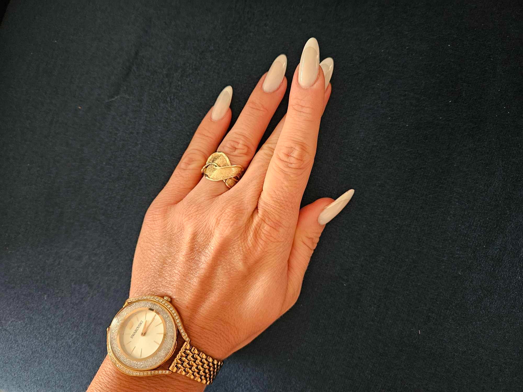 Dłoń z długimi, beżowymi paznokciami, złotym pierścionkiem i zegarkiem Swarovski na tle czarnego materiału.