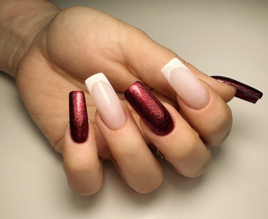 Dłoń z długimi paznokciami: dwa w odcieniach czerwieni z brokatem, dwa z białym french manicure i jeden bordowy na boku.