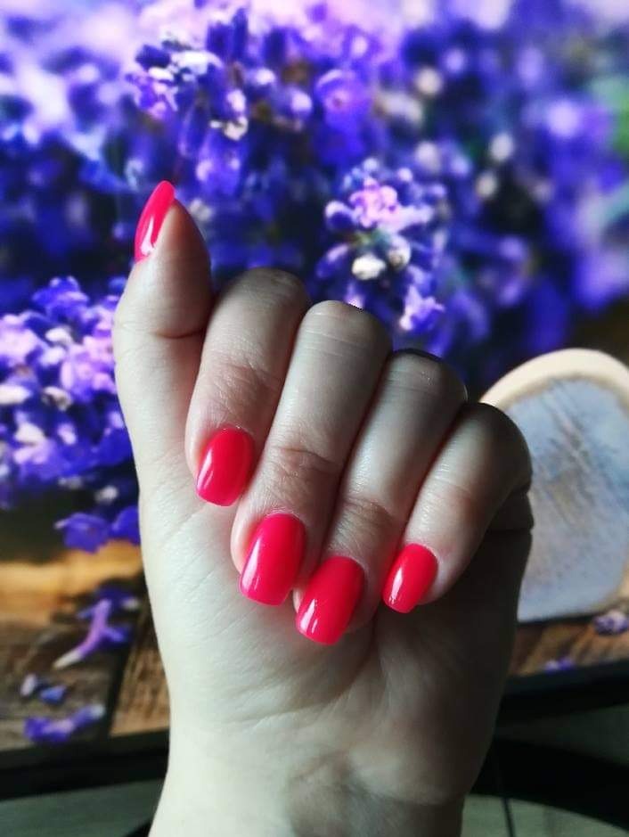 Dłoń z paznokciami pomalowanymi na intensywny, neonowy róż, na rozmytym tle z motywem lawendy.