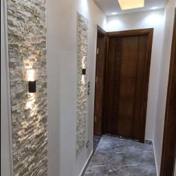 REMONTY I WYKOŃCZENIA PROFESIONALNIE-MATBUDPROFESIONAL MATEUSZ SIKORA - Wąski korytarz z dekoracyjną ścianą z kamienia, oświetleniem punktowym i marmurową podłogą prowadzący do drewnianych drzwi z geometrycznym wzorem.