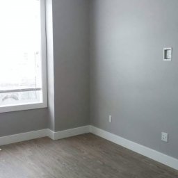 REMONTY I WYKOŃCZENIA PROFESIONALNIE-MATBUDPROFESIONAL MATEUSZ SIKORA - Pomalowane na szaro puste pomieszczenie z panelami podłogowymi, białymi listwami przypodłogowymi i oknem po lewej stronie.