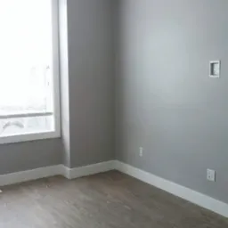 Pomalowane na szaro puste pomieszczenie z panelami podłogowymi, białymi listwami przypodłogowymi i oknem po lewej stronie.