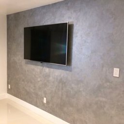 REMONTY I WYKOŃCZENIA PROFESIONALNIE-MATBUDPROFESIONAL MATEUSZ SIKORA - Ściana z efektem dekoracyjnym betonu architektonicznego w salonie, z zawieszonym telewizorem i gładką, białą podłogą.
