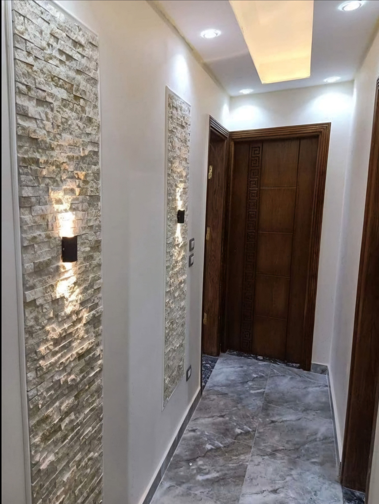 Wąski korytarz z dekoracyjną ścianą z kamienia, oświetleniem punktowym i marmurową podłogą prowadzący do drewnianych drzwi z geometrycznym wzorem.