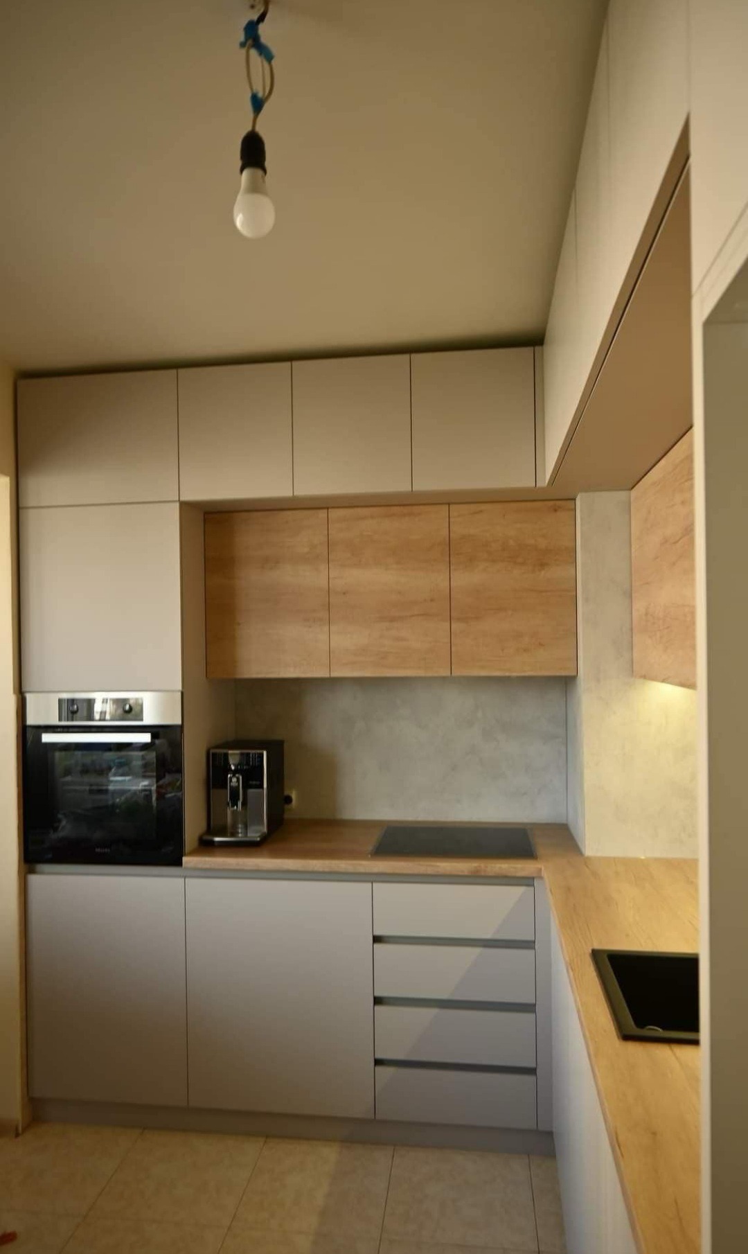 Nowoczesna kuchnia z szarymi szafkami, drewnianymi blatami i sprzętem AGD, z surową żarówką wiszącą na kablu.