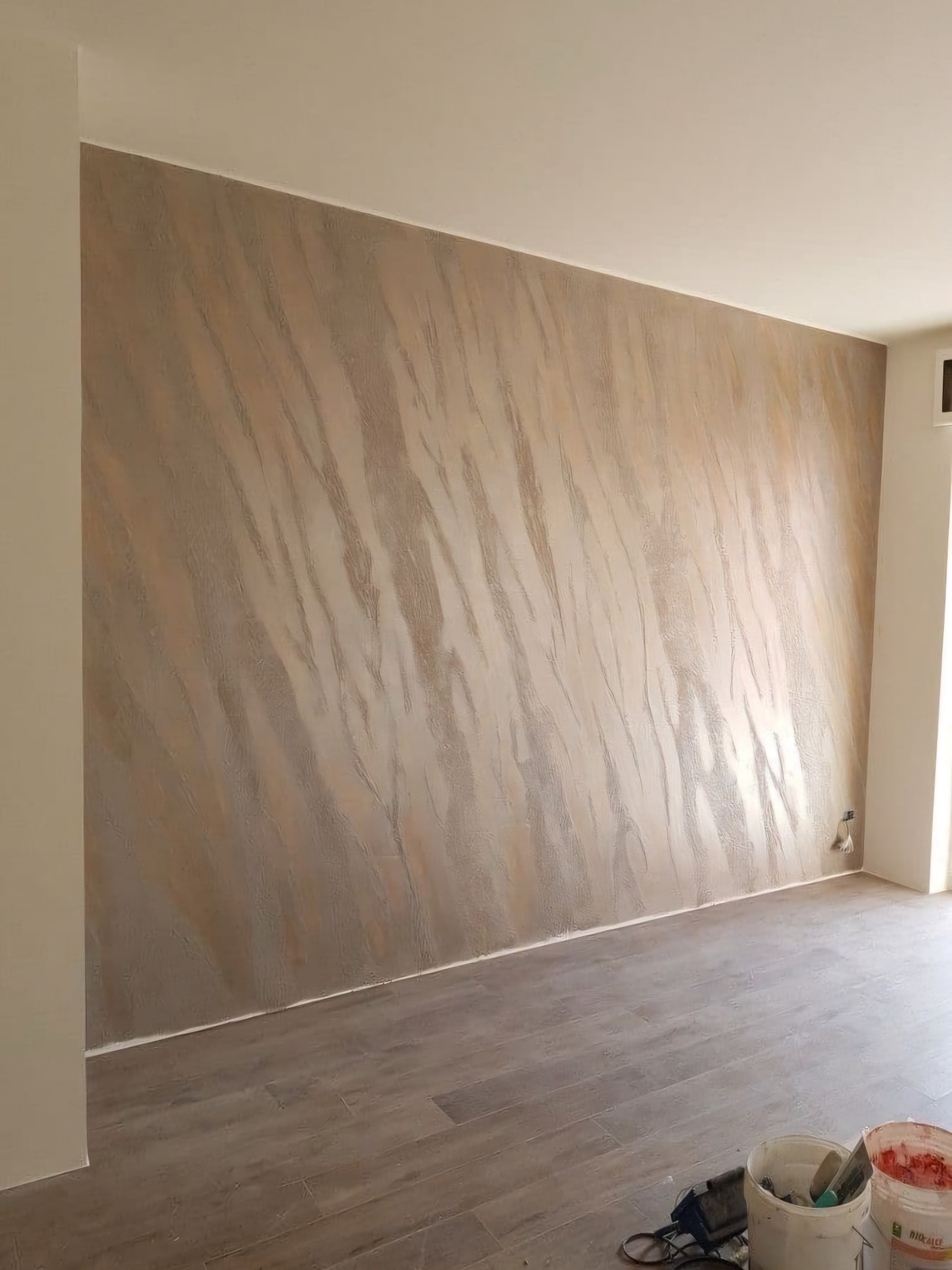 Ściana w pokoju po nałożeniu dekoracyjnej, beżowo-szarej gładzi imitującej marmurkowe żyły, widoczne wiaderka z narzędziami na podłodze.