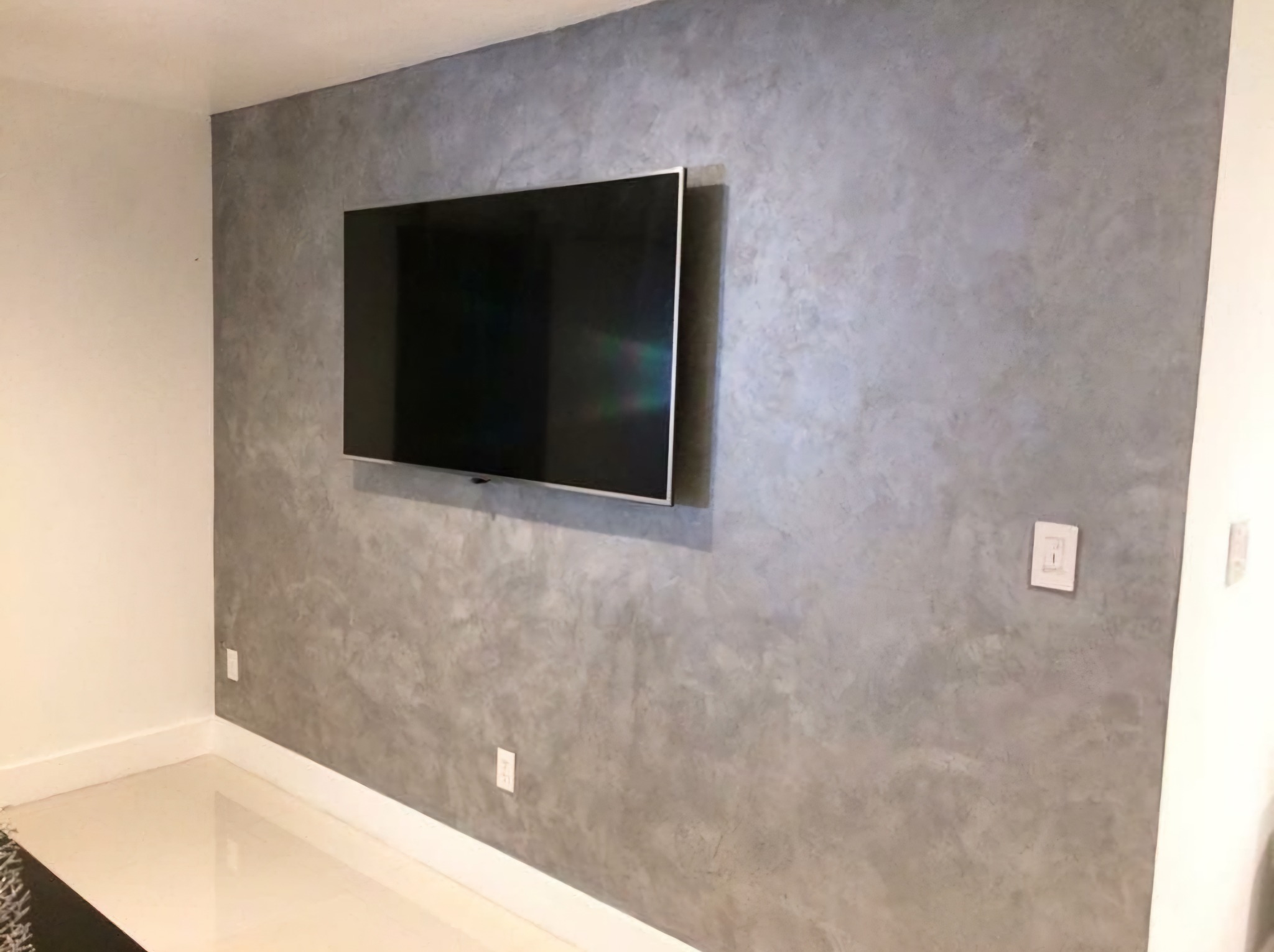 Ściana z efektem dekoracyjnym betonu architektonicznego w salonie, z zawieszonym telewizorem i gładką, białą podłogą.