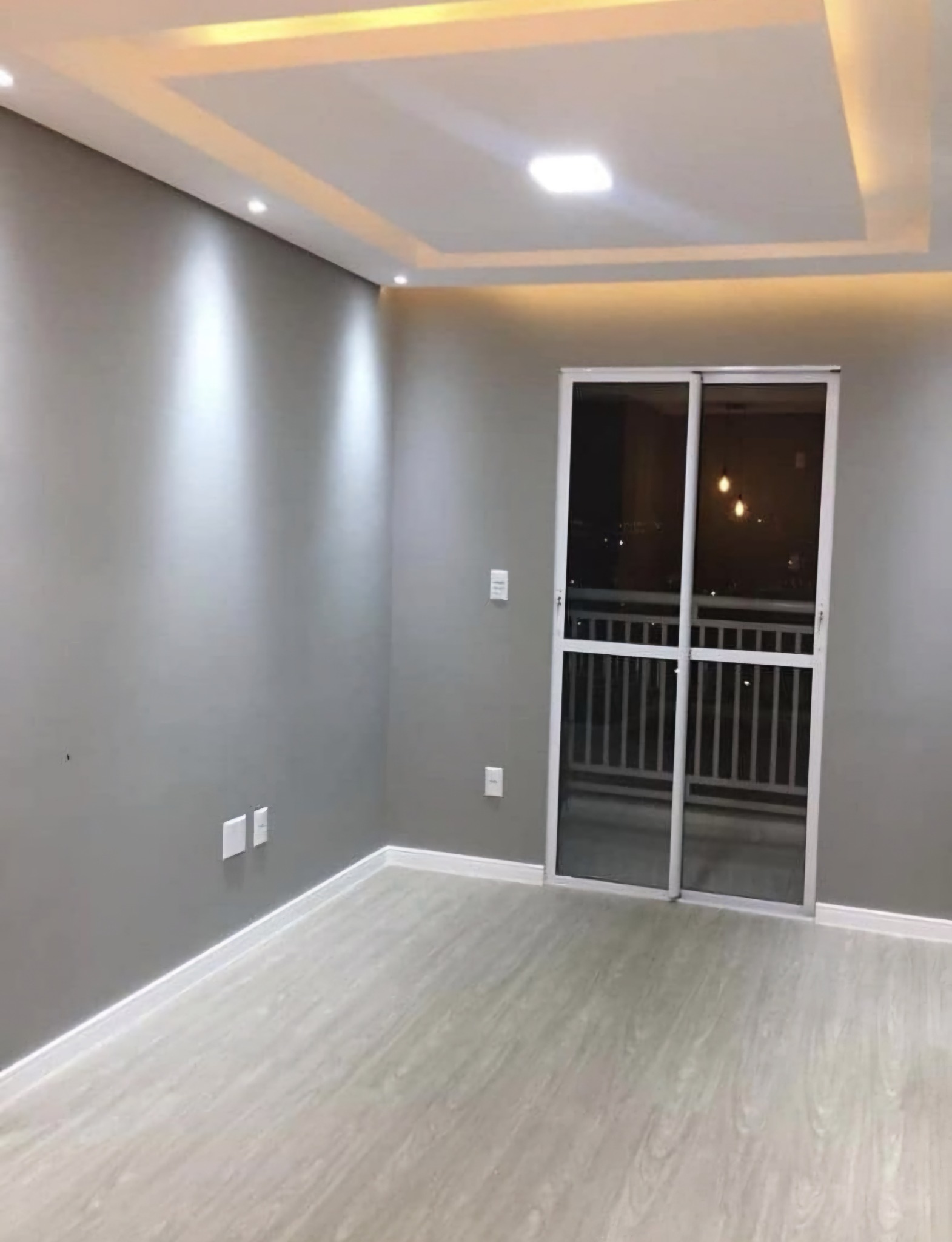 Wnętrze pokoju po odświeżeniu: szare ściany, jasna podłoga imitująca drewno, przesuwne drzwi balkonowe z widokiem na nocne miasto. Sufit podwieszany z oświetleniem punktowym i centralnym.