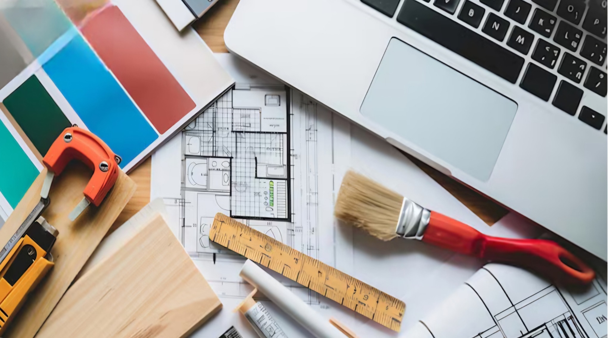 Paleta kolorów, plan mieszkania, pędzel z czerwoną rączką, laptop, miarka i szpachelki na stole.