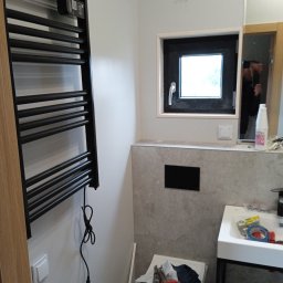 Adrian Korzeniewski - Wnętrze łazienki w trakcie remontu: widoczny czarny grzejnik drabinkowy z elektronicznym sterowaniem, okno z czarną ramą, częściowo wykończona ściana z szarą płytką, biała umywalka na metalowym...