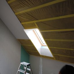 Adrian Korzeniewski - Montaż płyty karton gips na poddaszu z oknem dachowym, widoczna izolacja z żółtymi listwami montażowymi, drabina w tle.