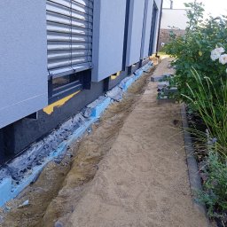Adrian Korzeniewski - Odsłonięty fundament budynku z widoczną hydroizolacją, zabezpieczony niebieskim styropianem, obok wykop z piaskiem i rosnące białe róże.