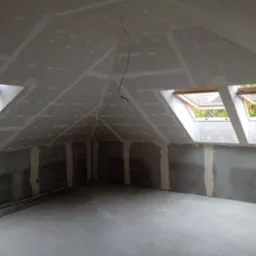 Poddasze w trakcie zabudowy płytami kartonowo-gipsowymi, widoczne szpachlowane łączenia, okna dachowe i wystający przewód elektryczny.