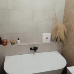 Nowoczesna łazienka z białą wanną, czarną armaturą i beżowymi płytkami, uzupełniona dekoracjami: suszone trawy w wazonie i pudełko prezentowe.