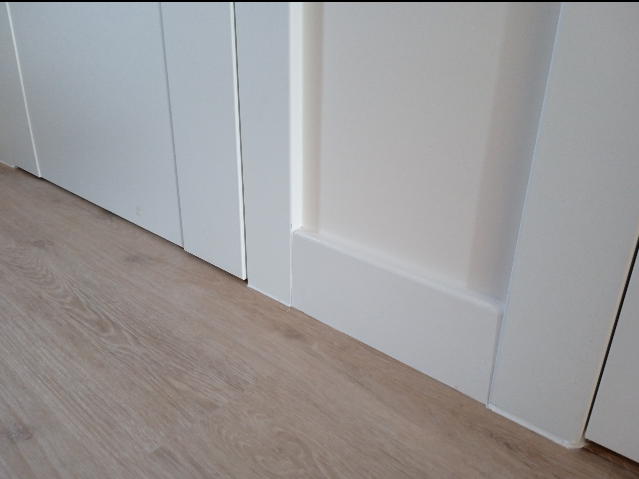 Detal wykończenia wnętrza: białe listwy przypodłogowe i panel podłogowy w odcieniach beżu, przy białej ścianie i drzwiach.