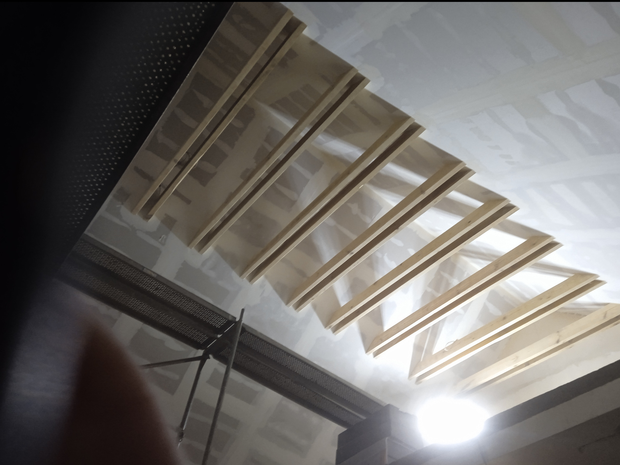 Szkielet konstrukcji z drewnianych belek pod zabudowę poddasza z płyt kartonowo-gipsowych, widoczny z dołu, z fragmentem rusztowania i oświetleniem budowlanym.