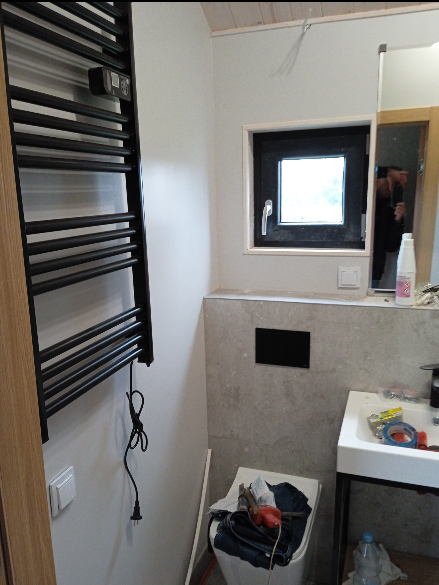 Wnętrze łazienki w trakcie remontu: widoczny czarny grzejnik drabinkowy z elektronicznym sterowaniem, okno z czarną ramą, częściowo wykończona ściana z szarą płytką, biała umywalka na metalowym...