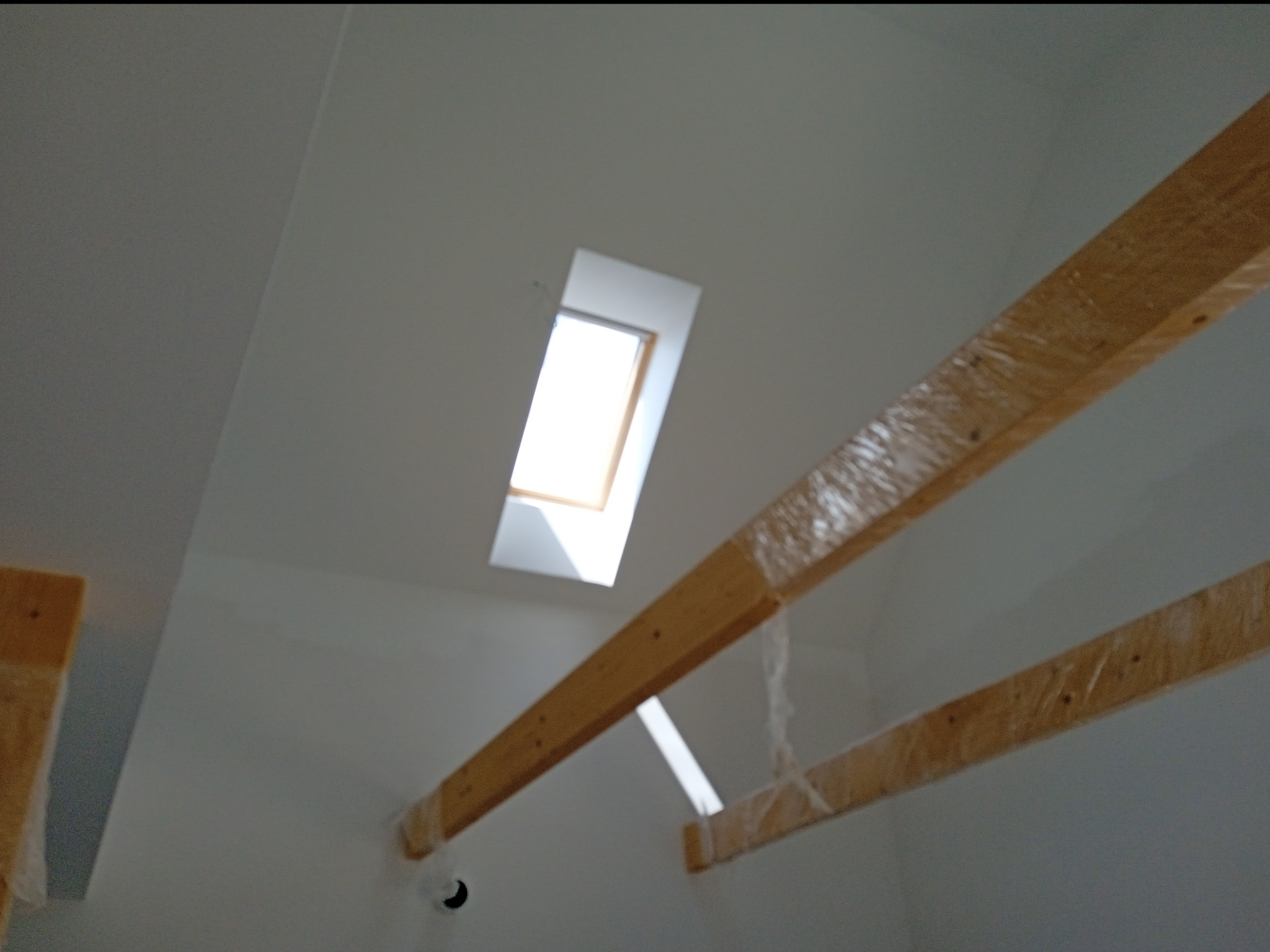 Sufit poddasza z oknem dachowym i elementami drewnianej konstrukcji zabezpieczonymi folią, przygotowany do dalszych prac wykończeniowych.