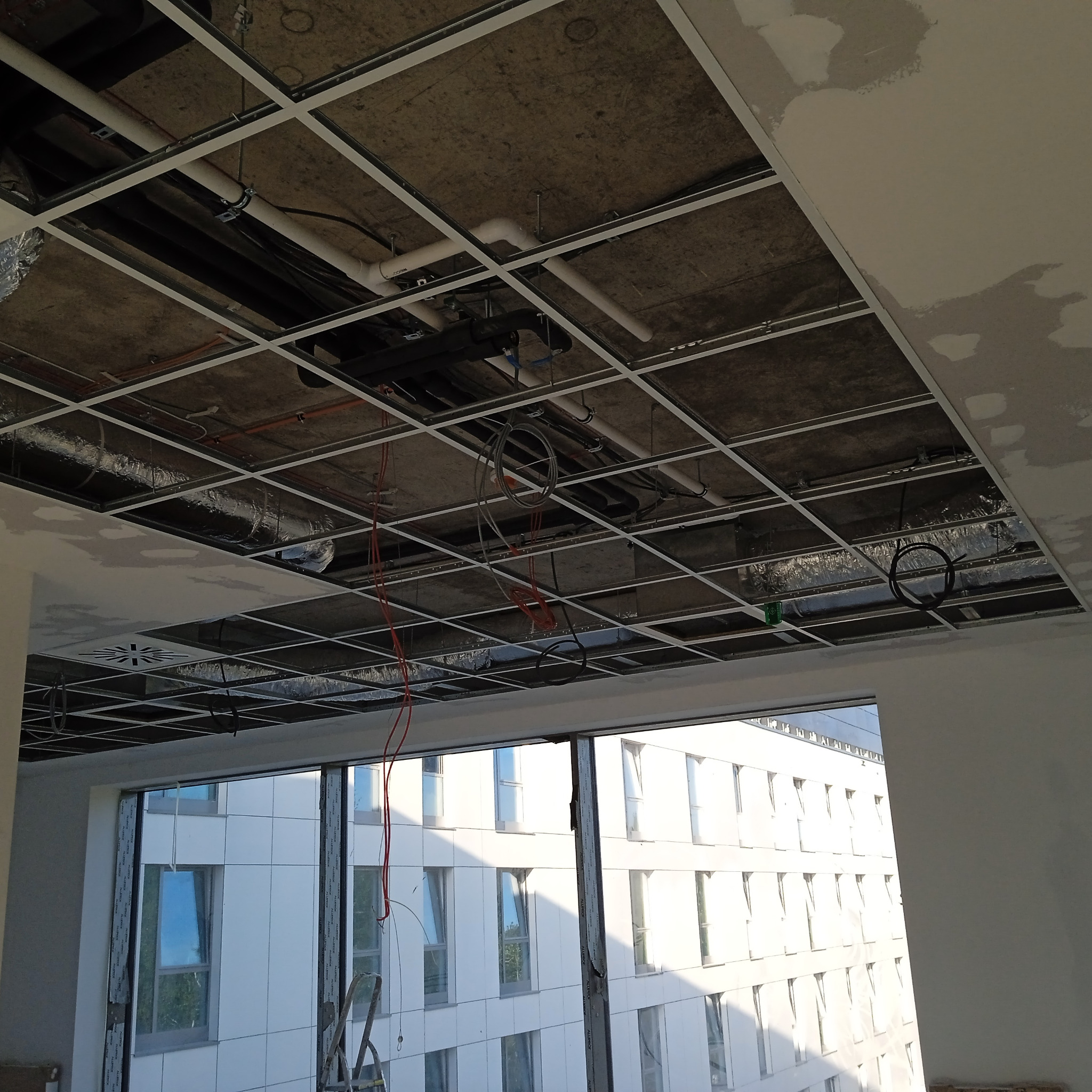 Montaż sufitu podwieszanego z widoczną konstrukcją metalową, instalacjami i przewodami w budynku szpitalnym, z widokiem na elewację budynku przez okno.