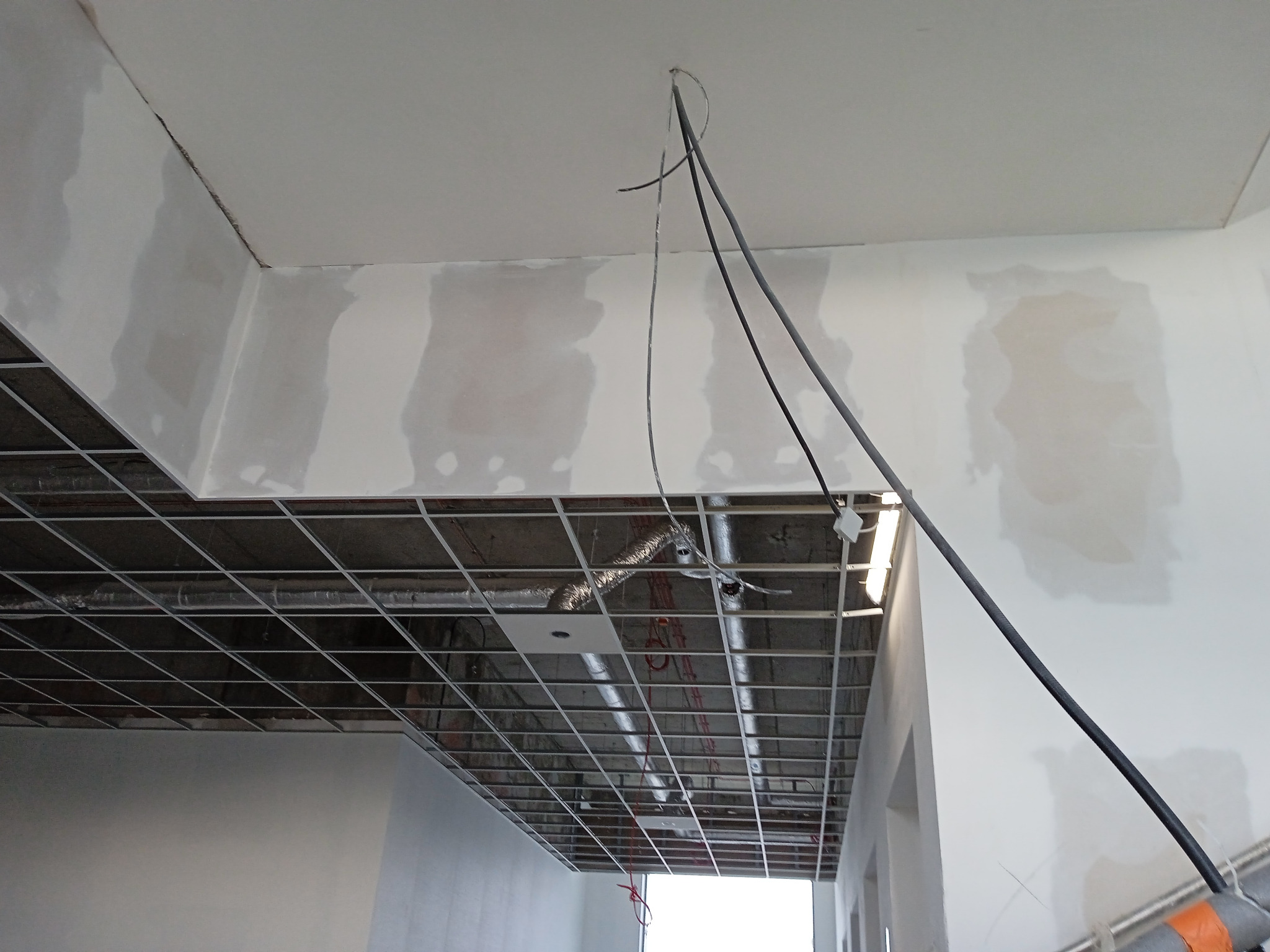Montaż sufitu podwieszanego z widoczną konstrukcją metalową, instalacjami i częściowo wykończonymi ścianami, na których widoczne są ślady szpachlowania.