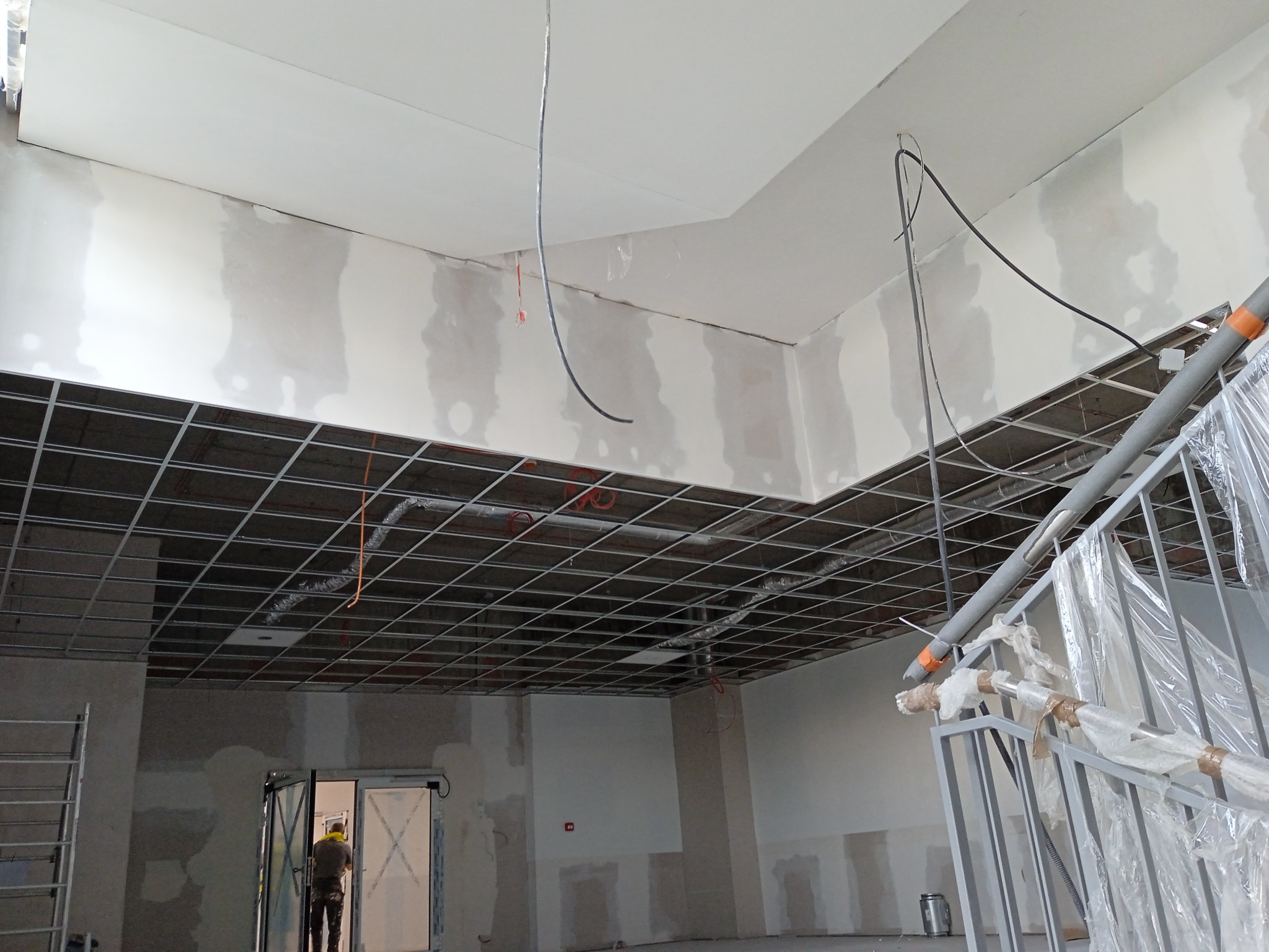 Sufit podwieszany z widoczną konstrukcją metalową w trakcie montażu w pomieszczeniu szpitalnym, częściowo wykończone ściany wymagające dalszych prac, widoczne przewody elektryczne.